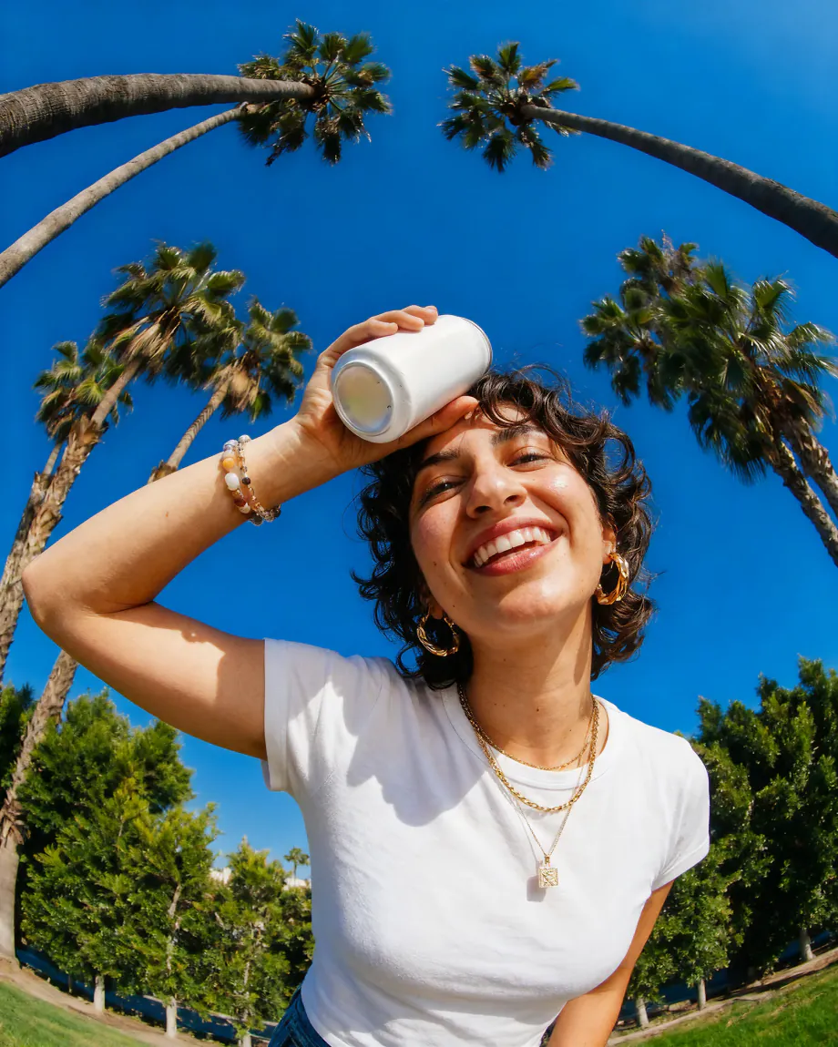 Woman in a white T-shirt laughing with a can pressed to her forehead under palm trees